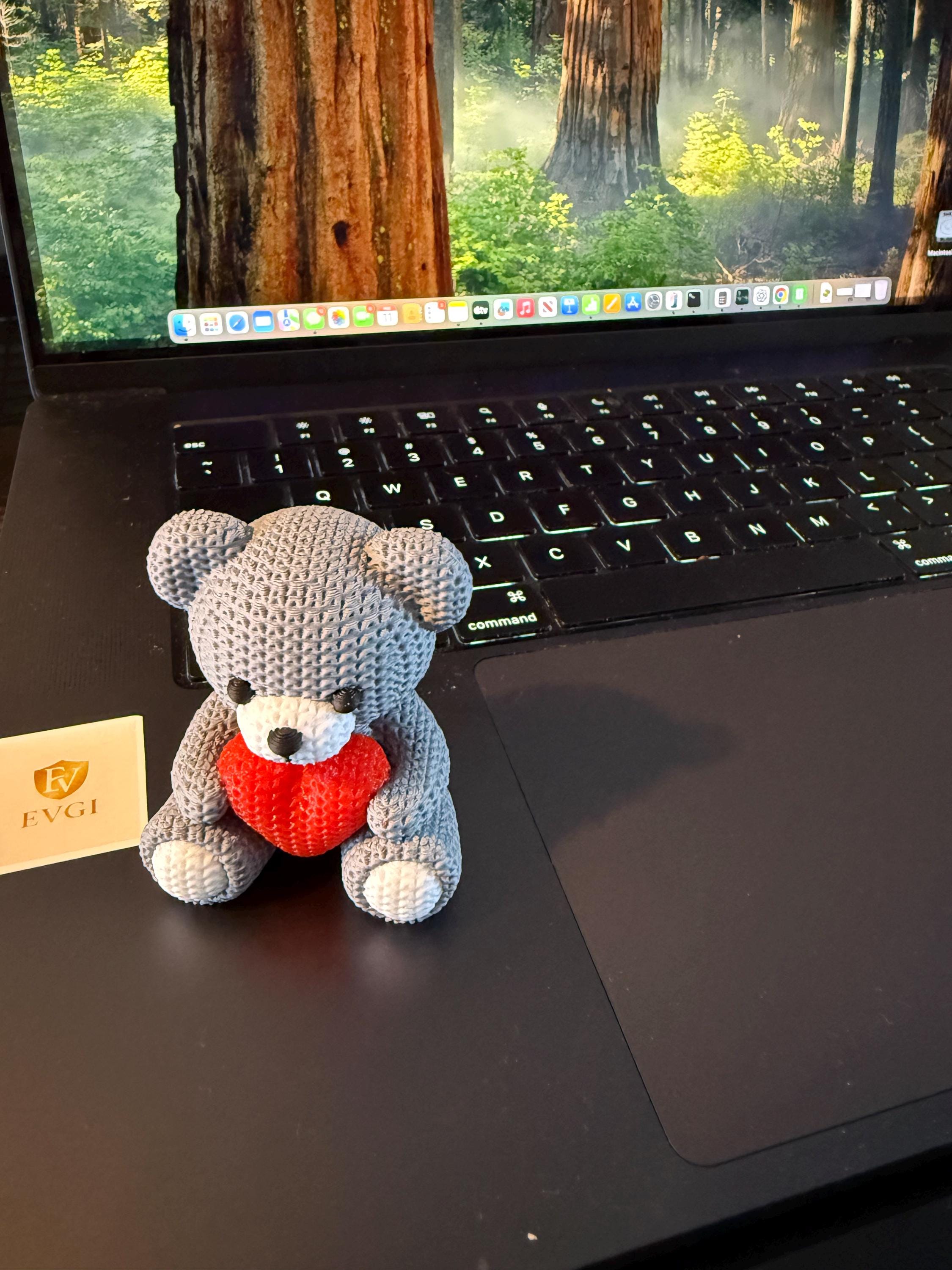 Crocheted teddy bear with a red heart on a desk in front of a laptop displaying a nature scene.